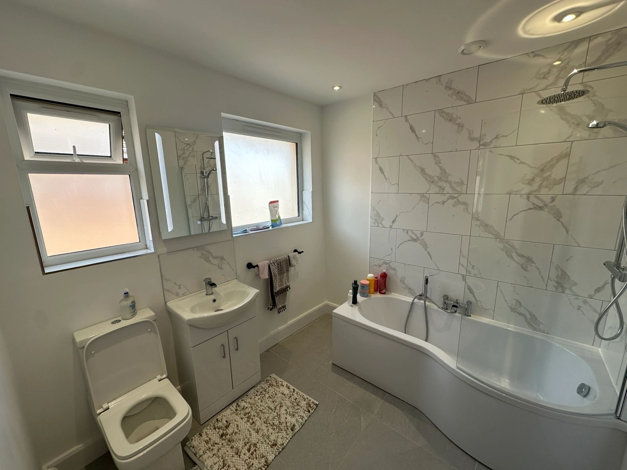 A finished bathroom with marble-effect tiling, a shower over a curved bath and a wall-hung basin.